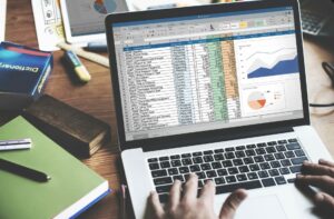 Vista desde arriba de una persona trabajando en una computadora portátil sobre un escritorio de madera. En la pantalla se muestra una hoja de cálculo con datos financieros detallados, un gráfico de líneas y un gráfico circular. El escritorio está rodeado de materiales de estudio como libros, una regla, marcadores y una tableta con gráficos adicionales.
