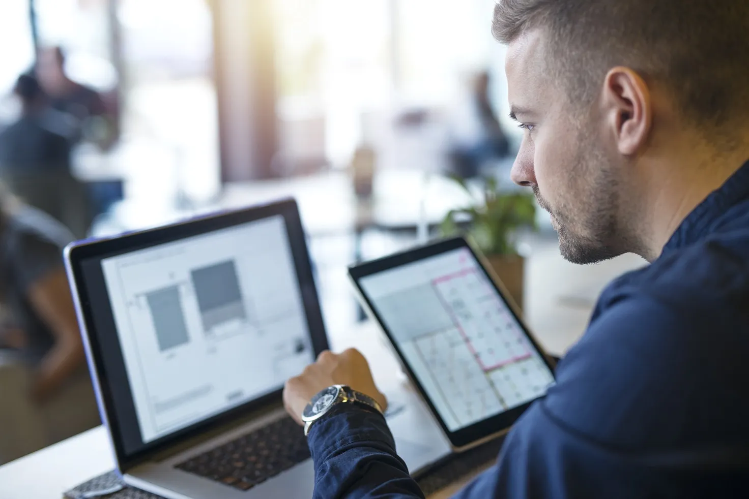 Hombre de perfil sosteniendo una tablet con datos y gráficos, mientras trabaja frente a una laptop abierta sobre un escritorio. Al fondo, una oficina luminosa y desenfocada con plantas y otra persona.