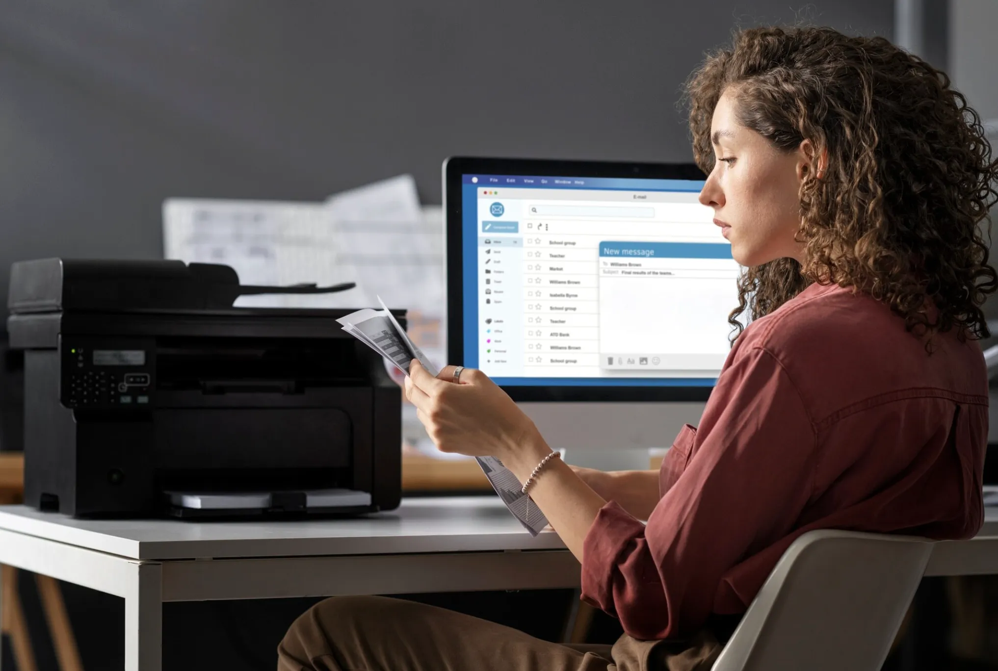 Mujer sentada en un escritorio revisando unos papeles con gráficos, frente a un monitor que muestra un cliente de correo electrónico con la bandeja de entrada y una ventana de mensaje nuevo. A la izquierda hay una impresora multifunción.